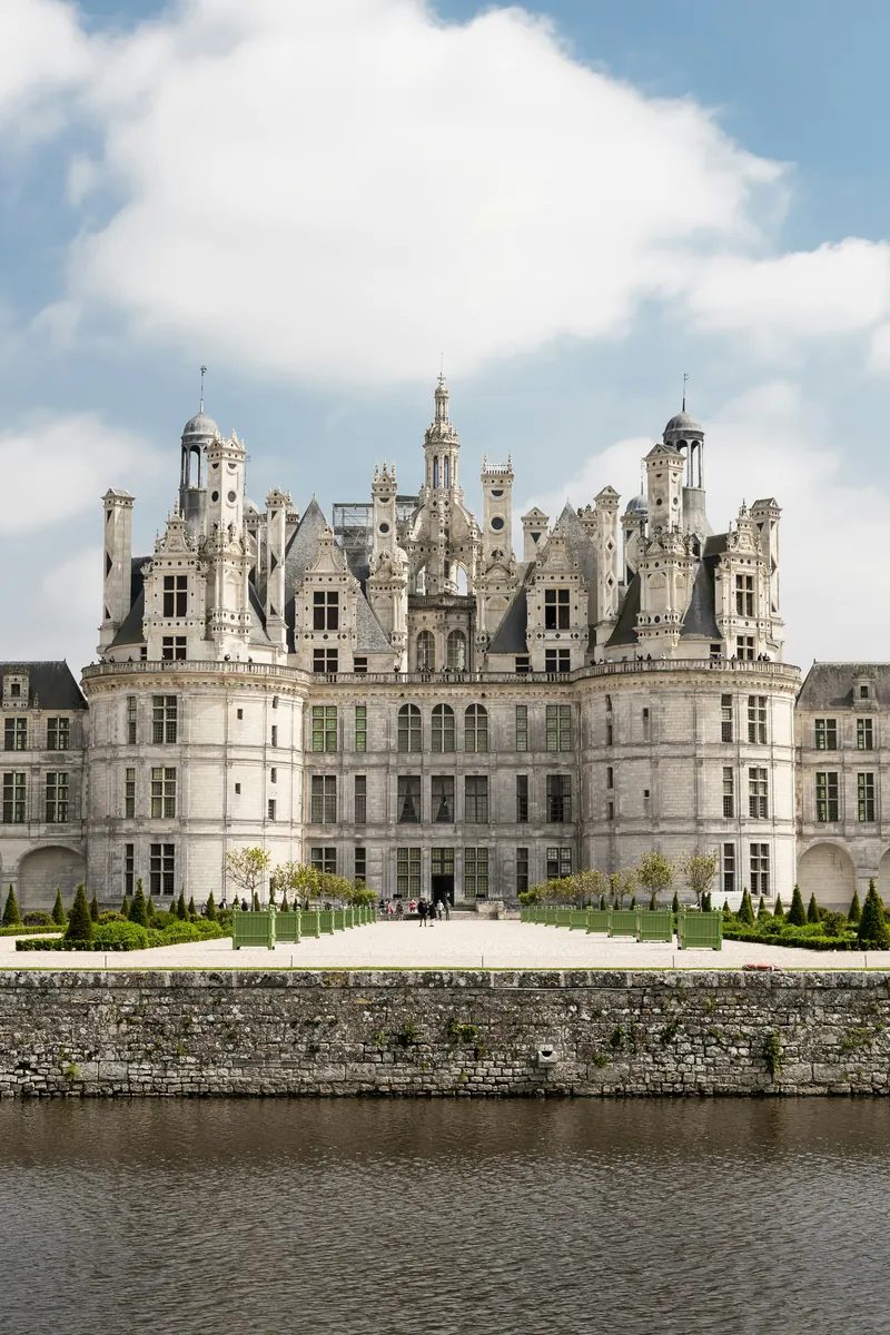 The Château de Chambord at golden hour — symmetrical white-stone keep with a fantastical roofline of turrets, lantern tower and hundreds of chimneys, reflected in the canal of its walled park. Loire Valley, France.
